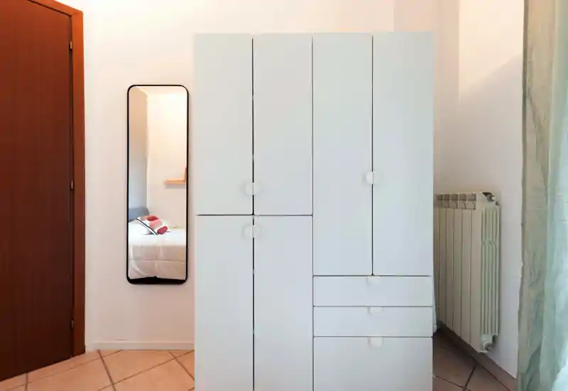 Vista di una camera da letto minimalista: un armadio bianco dominante e uno specchio a figura intera che riflette il letto con cuscini; luce naturale morbida e ambiente ordinato.