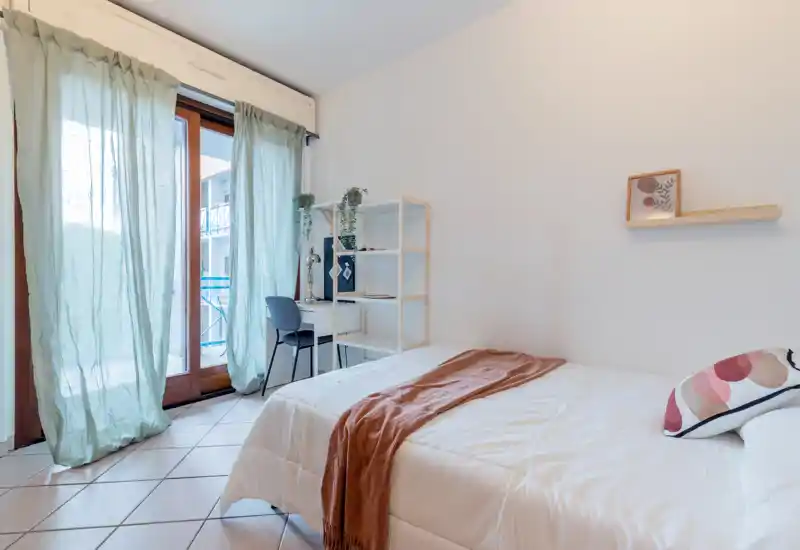 Dormitorio luminoso y acogedor con cama doble, manta y cojín decorativo, escritorio y estantería junto a la puerta del balcón que aporta abundante luz natural.