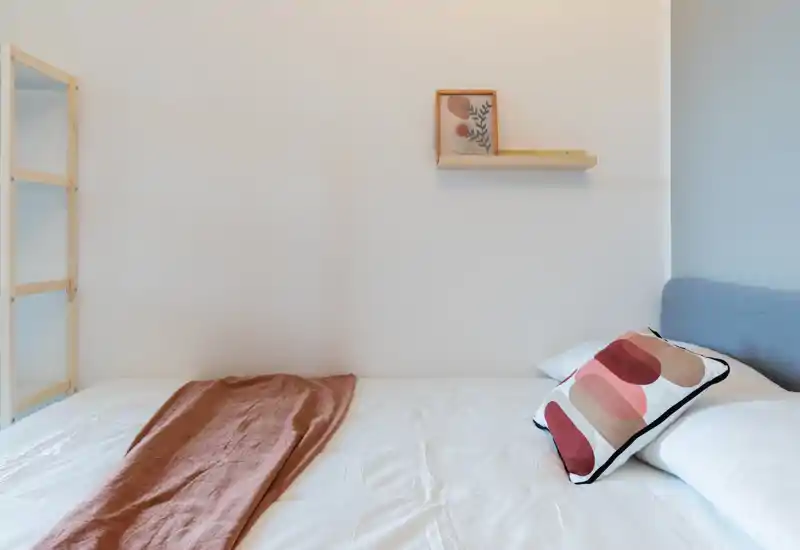 Dormitorio luminoso y minimalista con cama bien hecha, cojín decorativo y una manta cálida; paredes blancas y una estantería sencilla crean un ambiente moderno y sereno.