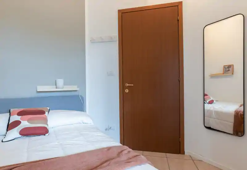 Dormitorio luminoso y ordenado con cama doble cómoda, cojín estampado y manta suave; paredes neutras y espejo de cuerpo entero que amplía el espacio.
