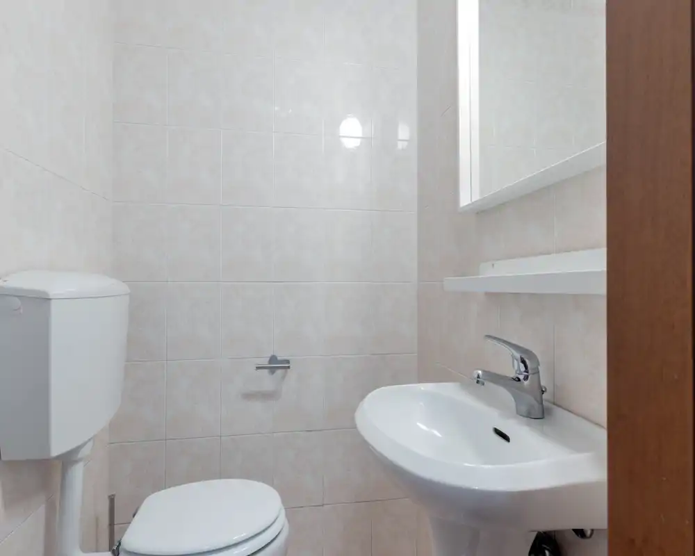 Compact, clean bathroom with a wall-mounted toilet, pedestal sink and mirror; neutral tiled walls and simple chrome fittings.