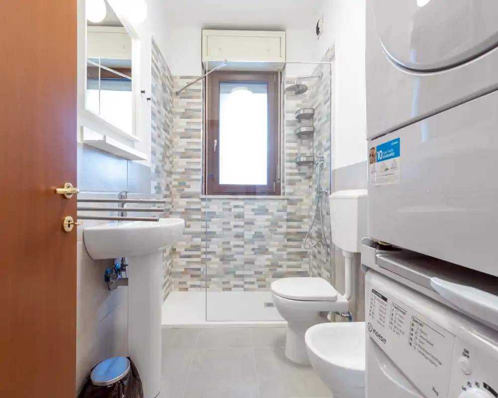 Clean, modern bathroom with walk-in shower, toilet, bidet and stacked washer/dryer unit. Neutral tiles and a window provide natural light.