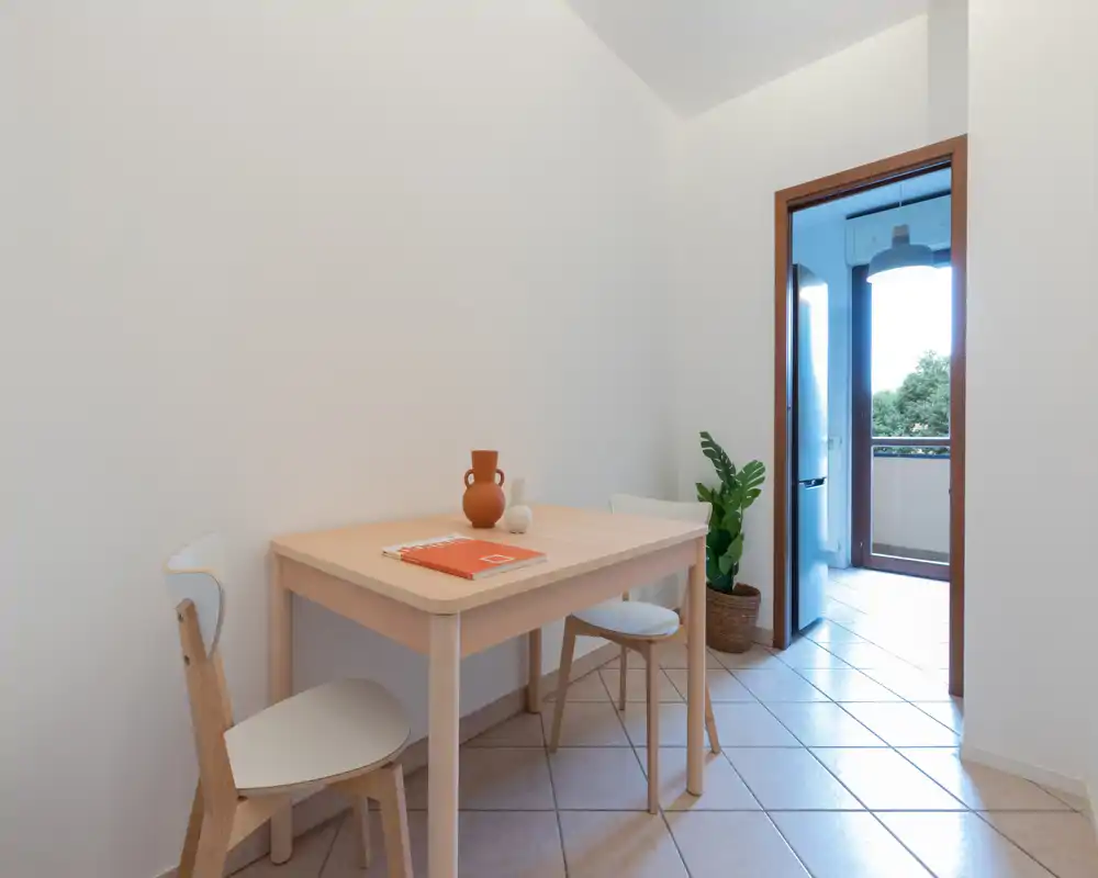 Small, bright dining corner with a light wood table, two chairs, a decorative vase and a potted plant; natural light pours in from an open doorway leading to a balcony.