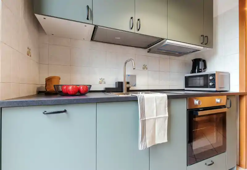 Cucina compatta e moderna con pensili verde menta, forno integrato, microonde e lavello con asciugamano. Ciotola decorativa e piano di lavoro ordinato creano un'area cucina accogliente.
