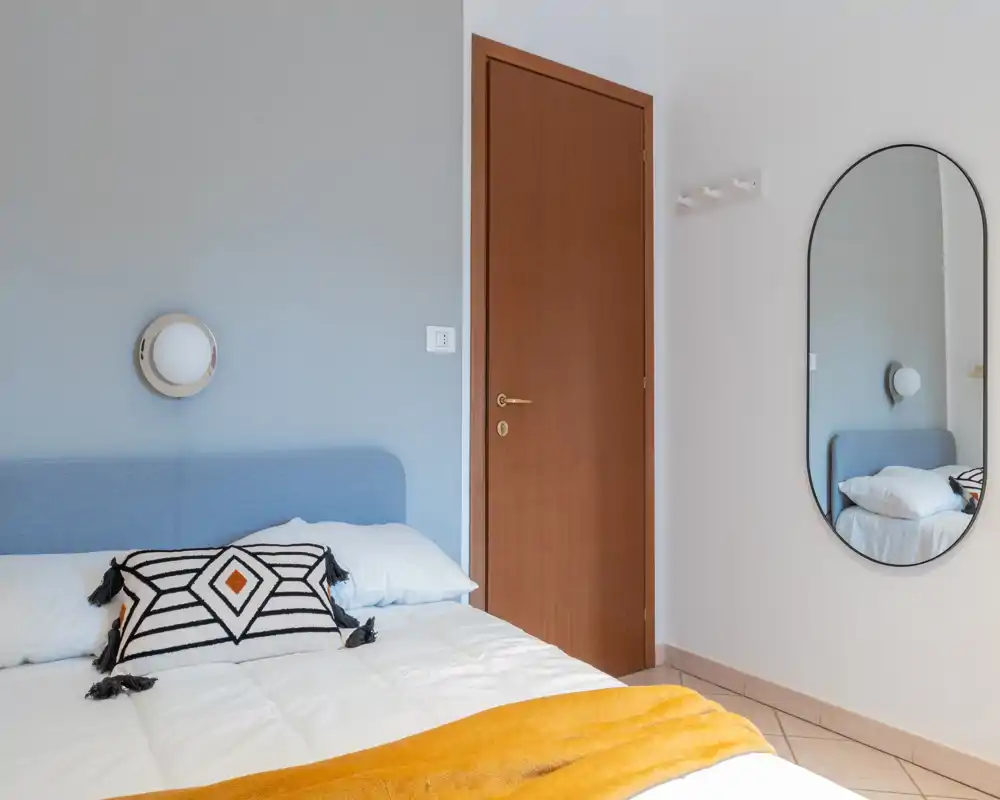 Bright, neatly styled bedroom with a blue upholstered headboard, decorative cushion and a warm yellow throw. The photo highlights a comfortable bed and simple modern details like a round wall light and oval mirror.