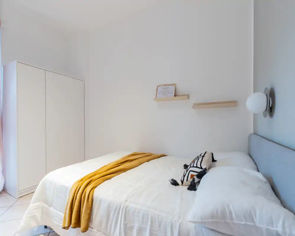 Bright, minimal bedroom with a neatly made double bed, decorative throw and cushion, and a freestanding wardrobe by the window.