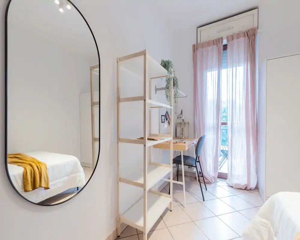 Bright, modern bedroom with soft pastel curtains, a simple desk by the balcony and a neatly made bed accented by a warm throw, offering a calm, airy atmosphere.