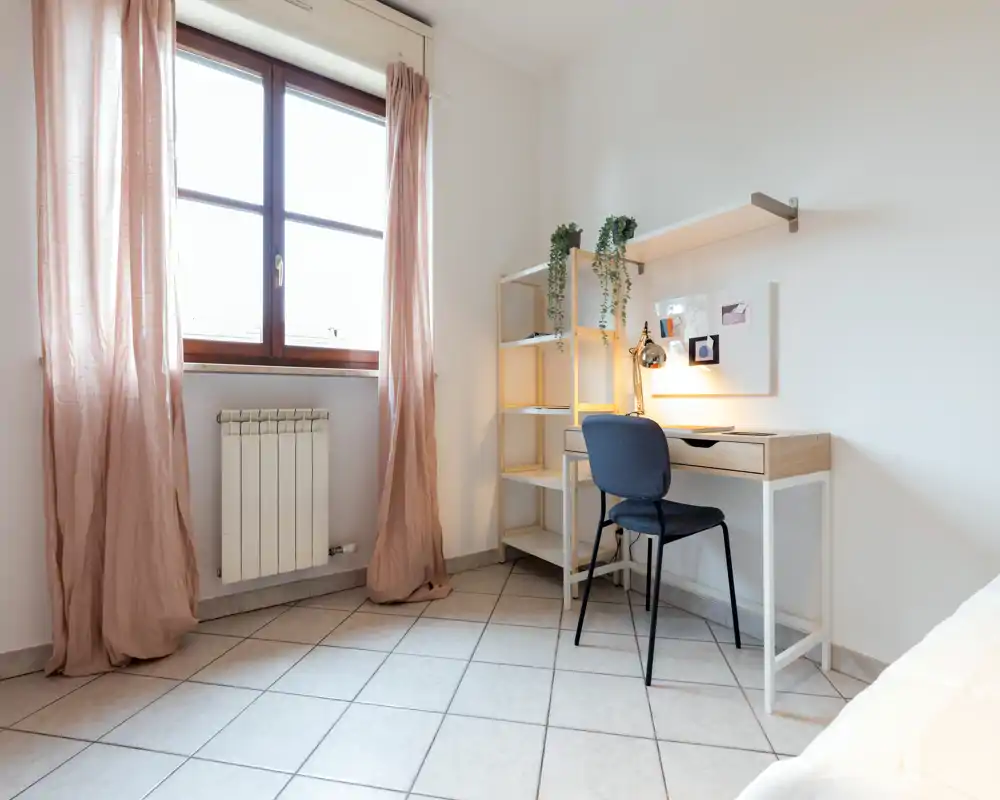 Compact bedroom corner with a small desk and shelving beside a window. Soft natural light, pale curtains and minimal decor create a bright, functional sleeping area.