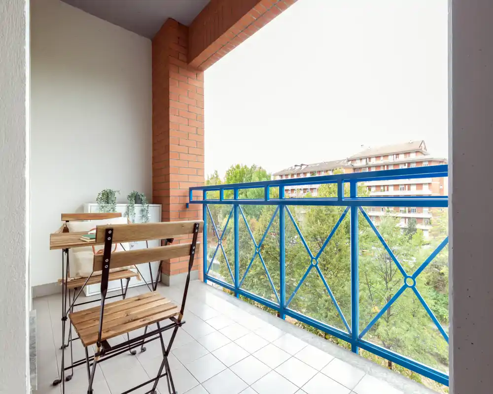 Sunny furnished balcony with a small wooden table and two folding chairs, blue metal railing and a pleasant view of trees and neighboring buildings.