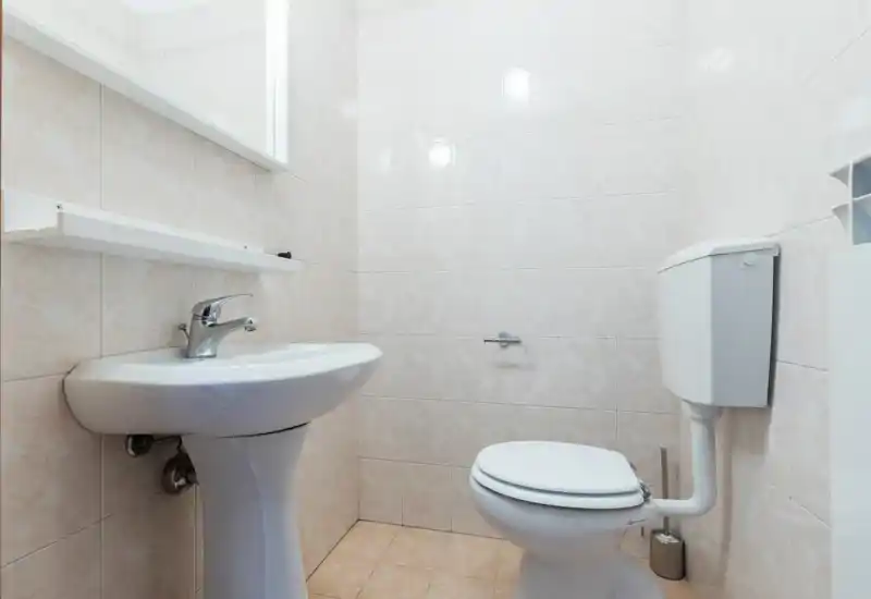 Compact, clean bathroom with a pedestal sink, wall-mounted mirror shelf and a standard toilet; light beige tiles and good lighting make the space feel tidy.