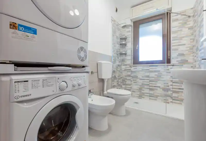 Modern bathroom with a shower enclosure, toilet, bidet and stacked washer/dryer; light tilework and a window provide bright, clean ambiance.