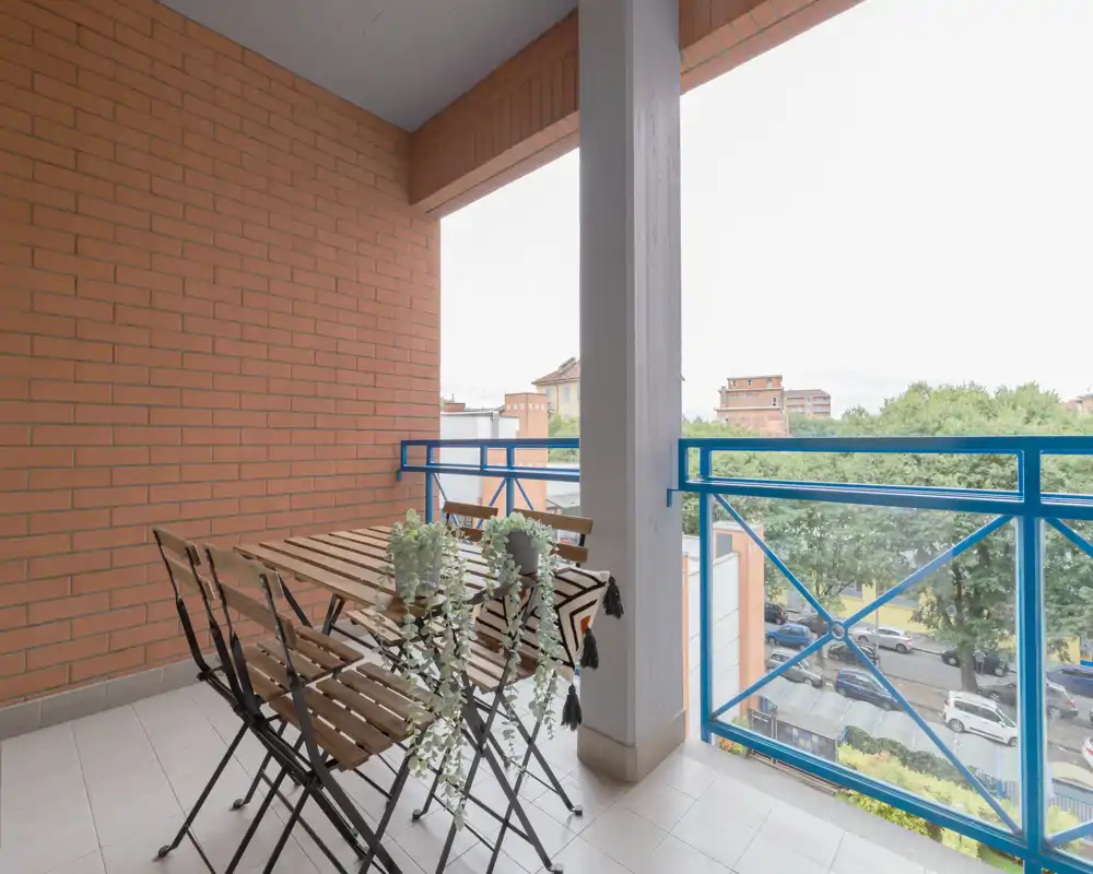 Balcón soleado con mesa de madera pequeña y sillas plegables, plantas decorativas y vistas a las copas de los árboles y la calle — ideal para el café matutino.