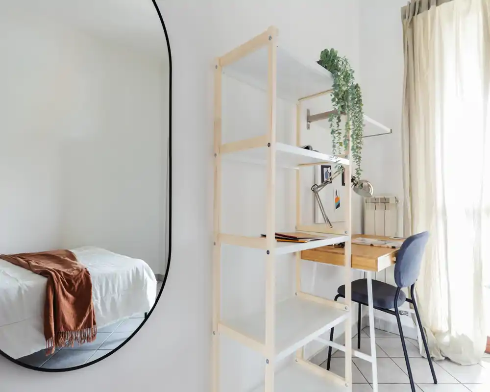 Rincón de dormitorio luminoso y minimalista con cama bien hecha, estantería de madera y un pequeño escritorio junto a una ventana soleada — elegante y funcional.