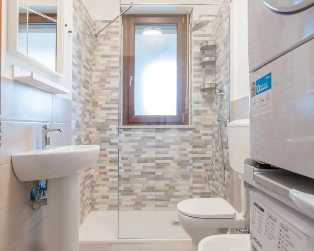 Baño moderno y luminoso con ducha a ras de suelo, ventana esmerilada para privacidad, lavabo de pedestal e inodoro; revestimiento tipo mosaico en tonos neutros y columna lavadora/secadora que aprovecha el espacio.