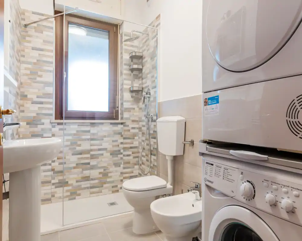 Baño moderno y luminoso con ducha a ras de suelo, inodoro, bidé y columna lavadora/secadora apilada — práctico y bien equipado.