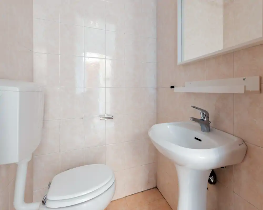 Baño pequeño y limpio con cisterna de inodoro montada en la pared, lavabo de pedestal y paredes alicatadas; luminoso y de acabado sencillo.