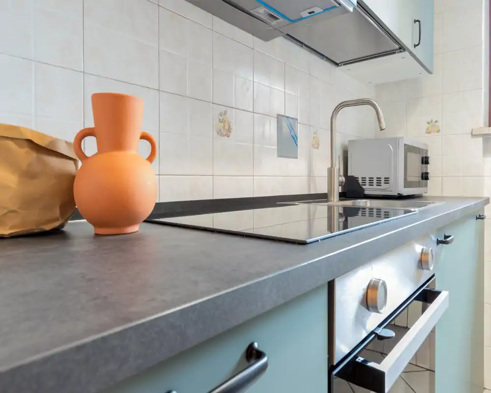 Angolo cucina compatto e moderno con piano cottura a induzione, forno incassato, miscelatore in acciaio e microonde sul piano. Piastrelle neutre e un vaso in terracotta aggiungono un tocco semplice e accogliente.