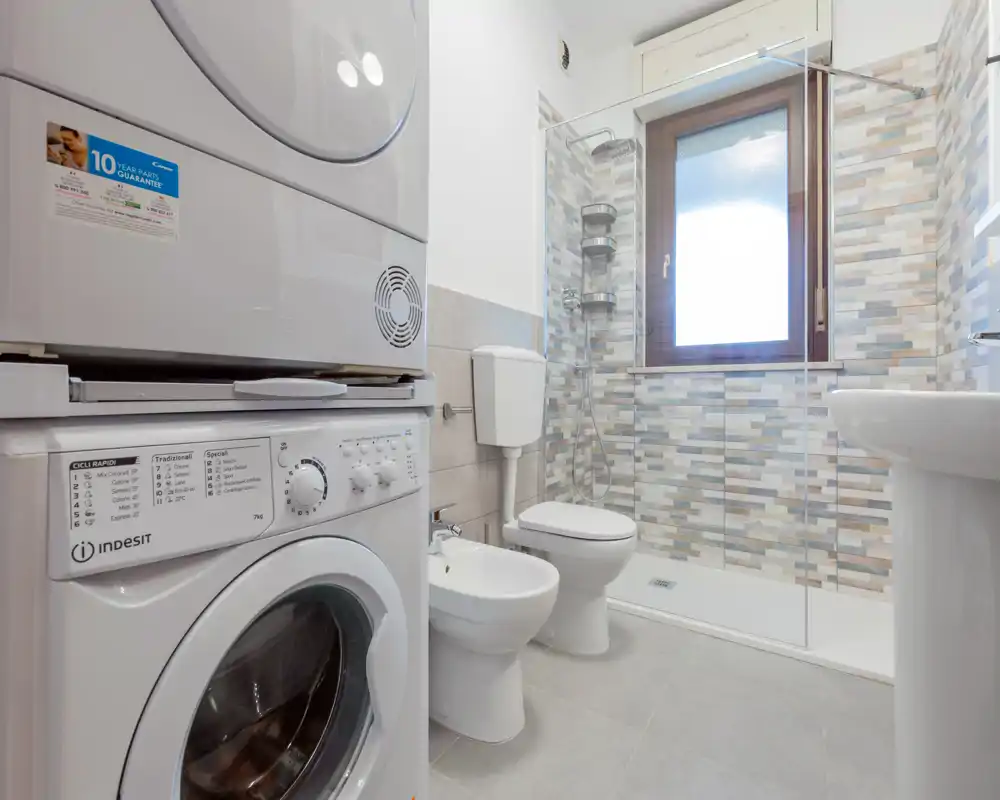 Bagno moderno e luminoso con doccia, wc, bidet e lavatrice/asciugatrice sovrapposte. Piastrelle pulite e una grande finestra che garantisce ottima luce naturale.