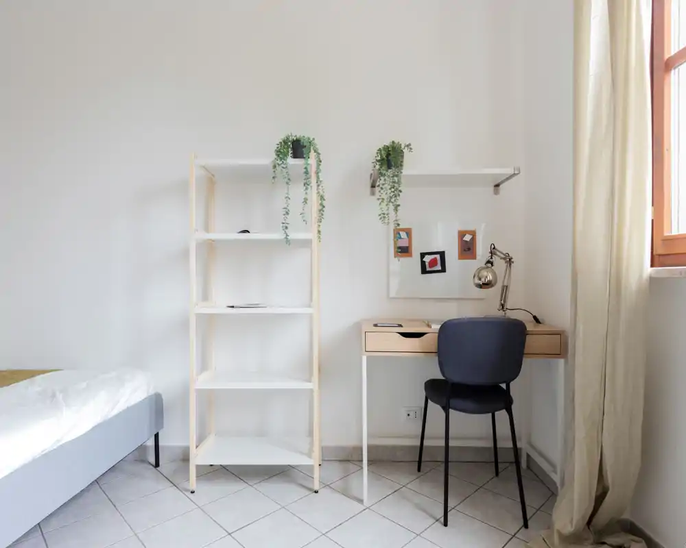 Angolo camera luminoso e minimale con bordo del letto visibile, scaffale aperto e una scrivania in legno vicino alla finestra; ideale per una persona o uno studente.