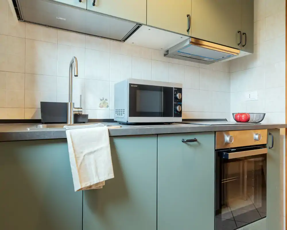 Cocina moderna y compacta con armarios verdes, horno empotrado, microondas sobre la encimera, fregadero de acero y paño; bien iluminada y presentada para anuncios.