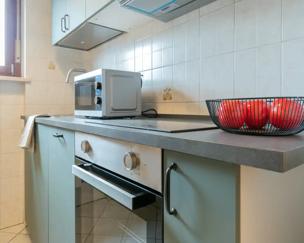 Encimera de cocina luminosa y ordenada con horno, placa de inducción, microondas y un cuenco decorativo con tomates; azulejos neutros y ventana que aportan luz natural.