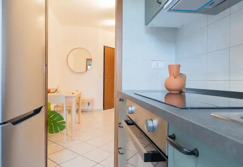 Modern compact kitchen with an induction cooktop, built-in oven and refrigerator visible in the frame; light tile backsplash and neutral countertops create a clean, bright cooking area.
