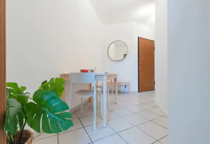 Bright, minimal entrance area with a small table and chair, round mirror and tiled floor — functional and welcoming for arrivals.