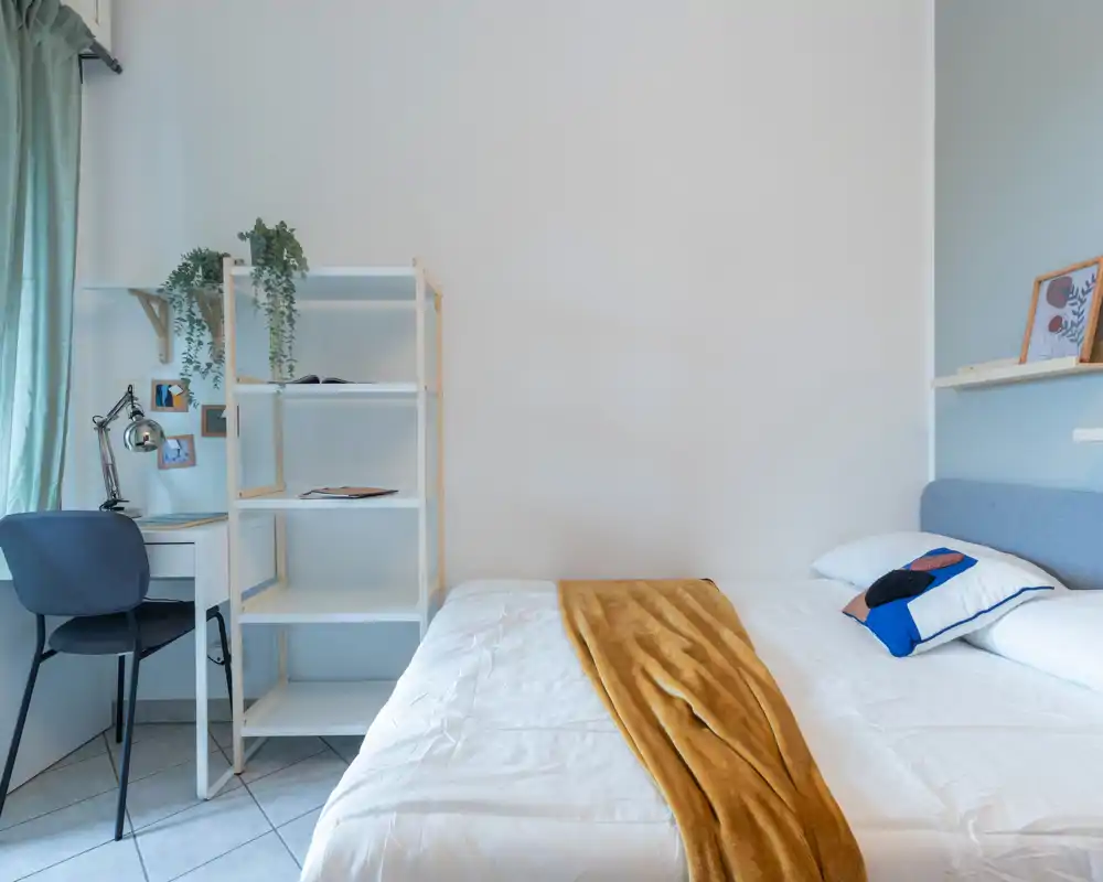 Chambre lumineuse et minimaliste avec un lit double bien fait, un jeté doux et un coussin décoratif ; un petit bureau et des étagères forment un espace de travail près de la fenêtre.