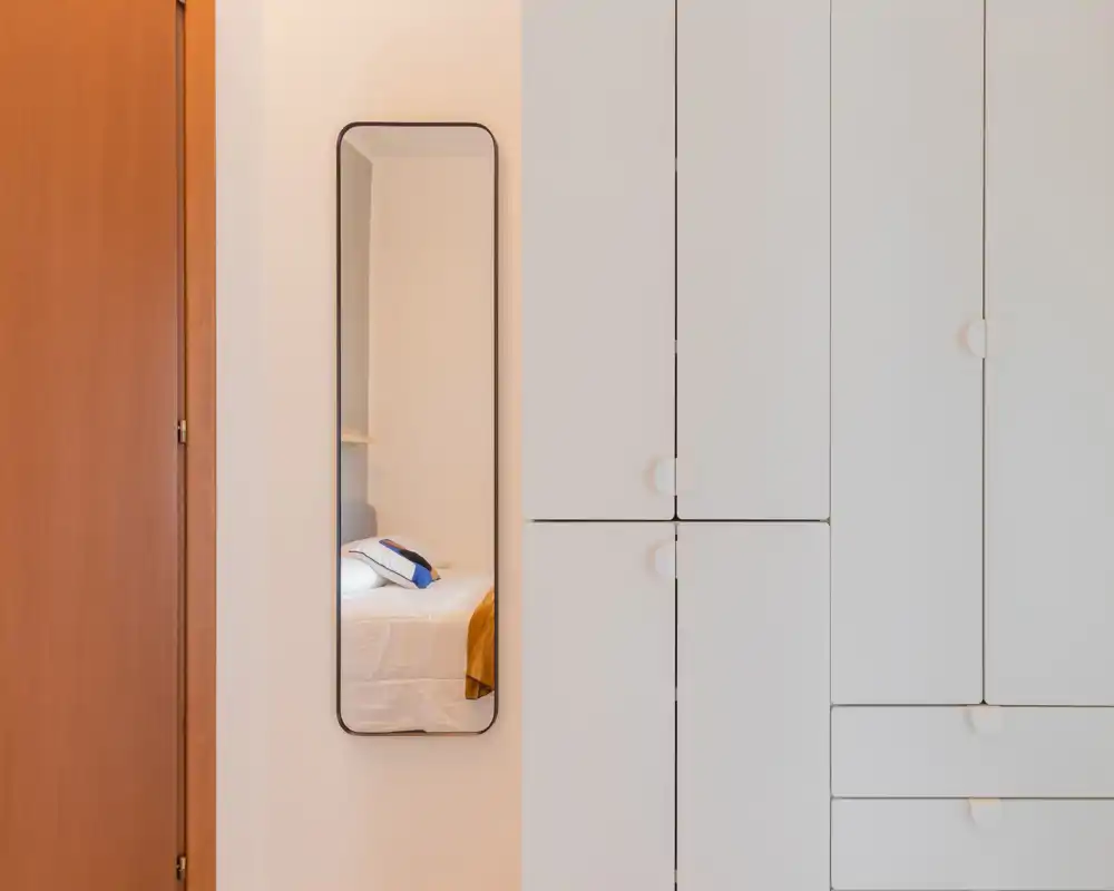 Un aperçu de la chambre vu dans un miroir mural étroit : un lit fait avec oreillers et plaid en reflet, des armoires blanches encastrées et une porte en bois au premier plan.