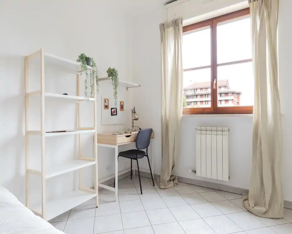 Coin chambre lumineux et minimaliste avec un petit bureau, des étagères et une fenêtre laissant entrer beaucoup de lumière naturelle.