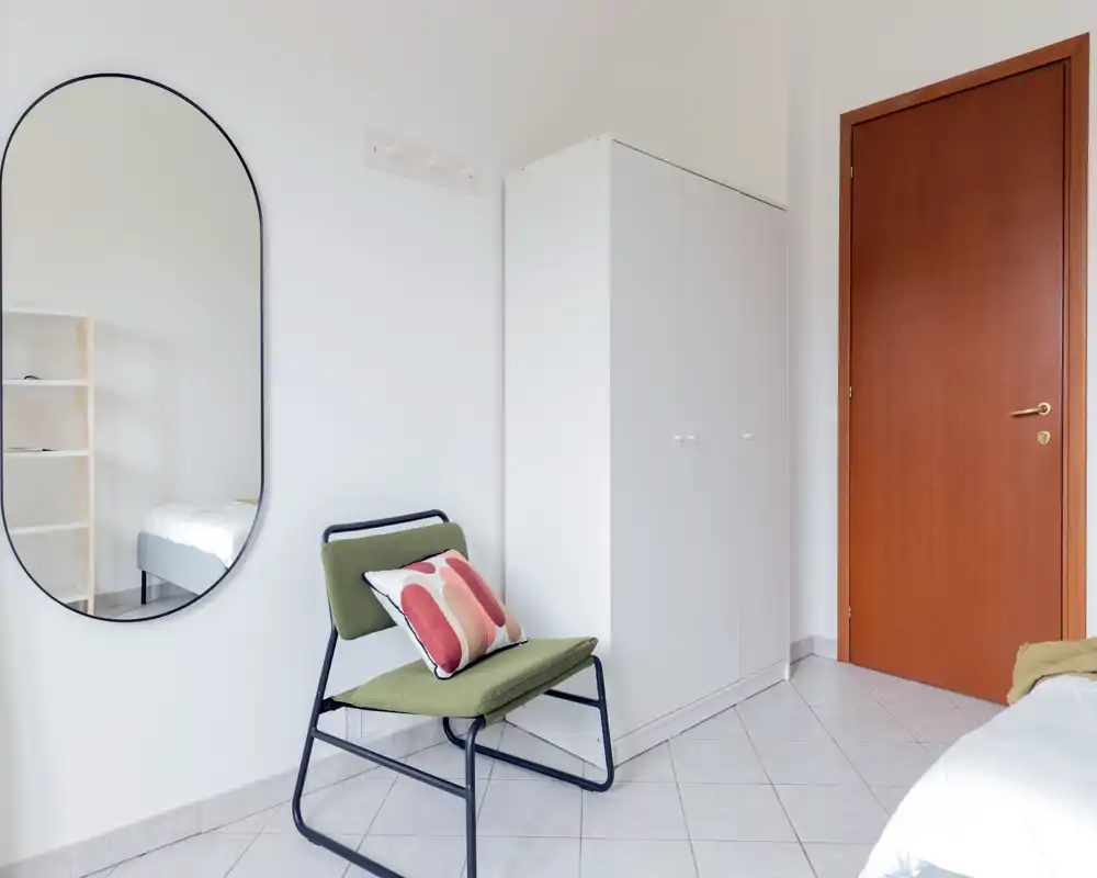 Coin de chambre lumineux et minimaliste avec un fauteuil moderne, coussin décoratif, armoire et vue partielle du lit ; murs blancs et grand miroir pour plus de profondeur.