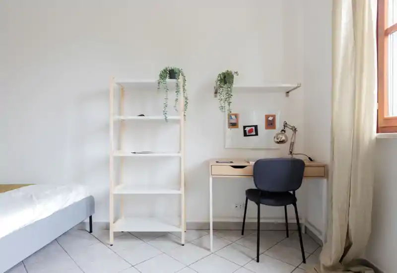 Coin chambre minimaliste et lumineux avec un lit rangé, une étagère ouverte et un petit bureau en bois près de la fenêtre avec chaise et lampe. Des plantes tombantes et des textiles neutres apportent une ambiance calme et moderne, idéale pour une annonce locative.