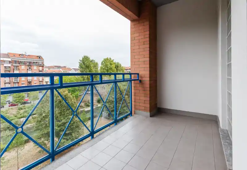 Balcon couvert avec sol carrelé et garde-corps métallique bleu décoratif, donnant sur des arbres et des immeubles en brique — parfait pour un coin repas ou détente.