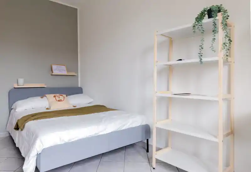 Chambre lumineuse et minimaliste avec un lit double bien fait, cadre rembourré gris clair, étagères simples et plante décorative ; ambiance calme et aérée, parfaite pour une annonce.