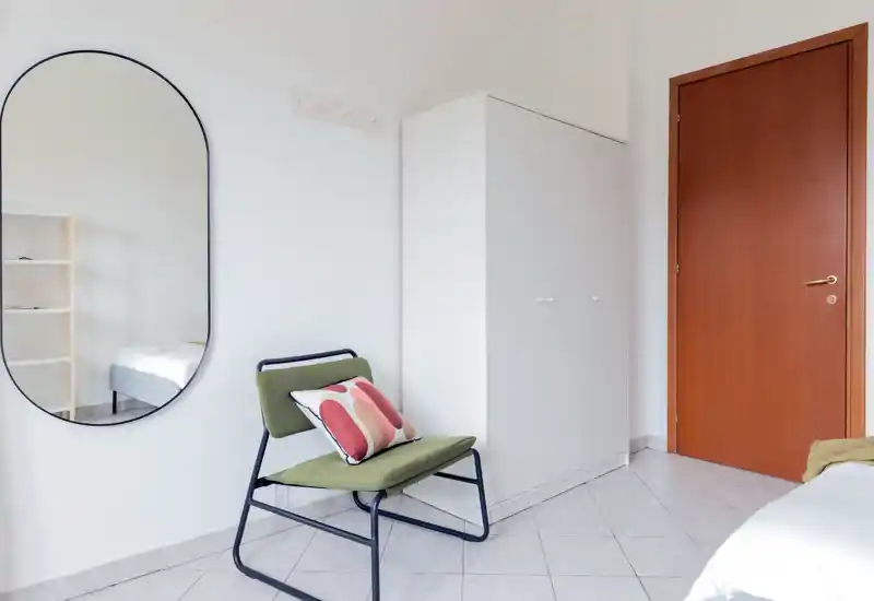 Coin de chambre lumineux et minimaliste avec un fauteuil moderne, coussin décoratif, armoire et vue partielle du lit ; murs blancs et grand miroir pour plus de profondeur.