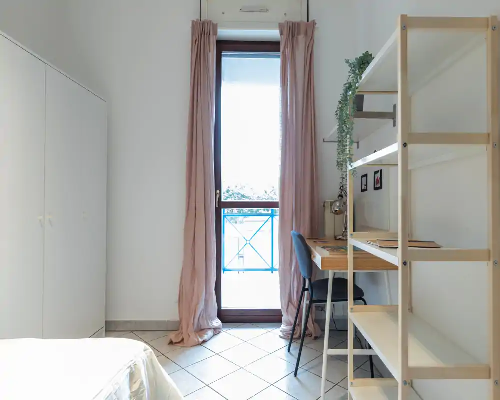 Chambre lumineuse et minimaliste avec un petit bureau et des étagères à côté d'une porte-fenêtre. La photo met en valeur la lumière naturelle, un mobilier simple et des rideaux doux pour une ambiance apaisante.