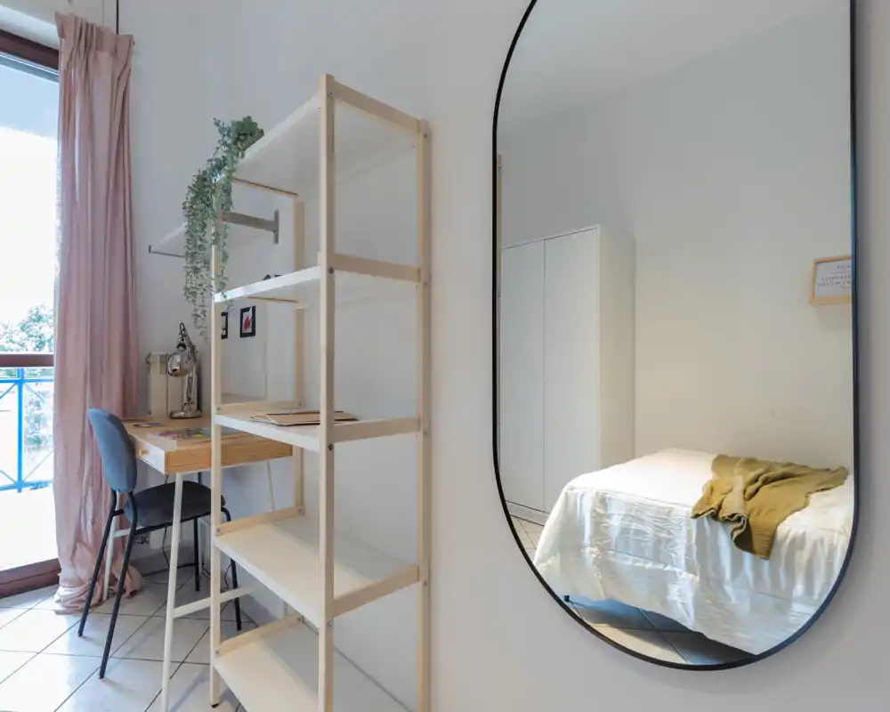 Coin chambre lumineux et minimaliste avec étagère en bois clair, un petit bureau près de la fenêtre et le lit visible dans le miroir; décoration épurée et lumineuse avec des textiles doux.