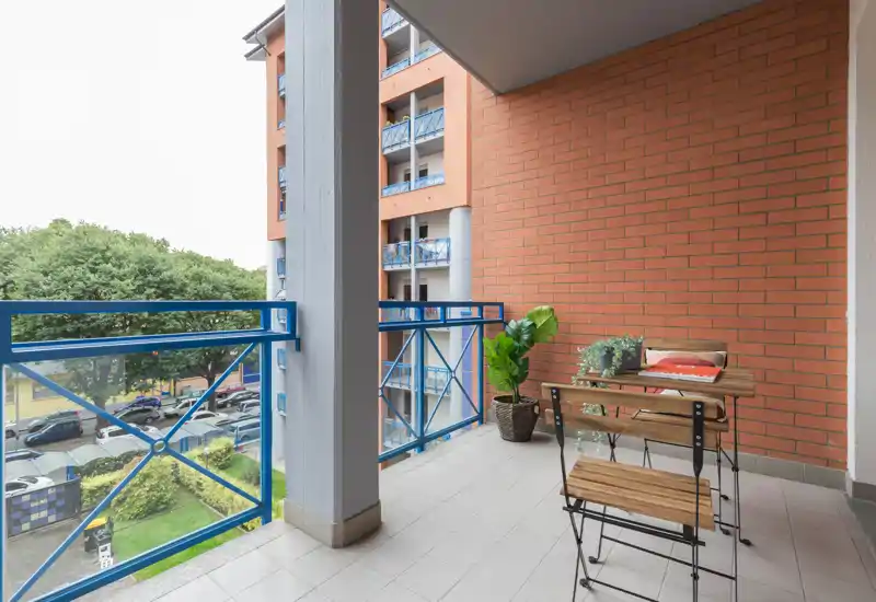 Sunny furnished balcony with a small wooden table and two chairs, decorative plants and a clear view over trees and the courtyard.