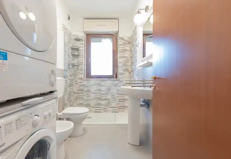 Bright, modern bathroom with walk-in shower, pedestal sink, toilet and stacked washer-dryer. Neutral mosaic tiles behind the shower and a window provide natural light.