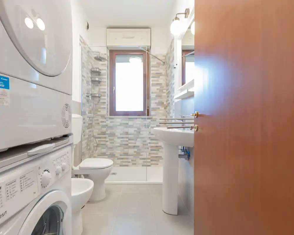 Bagno moderno e luminoso con doccia walk-in, WC e lavabo; parete con piastrelle a mosaico e lavatrice/asciugatrice impilate a sinistra.