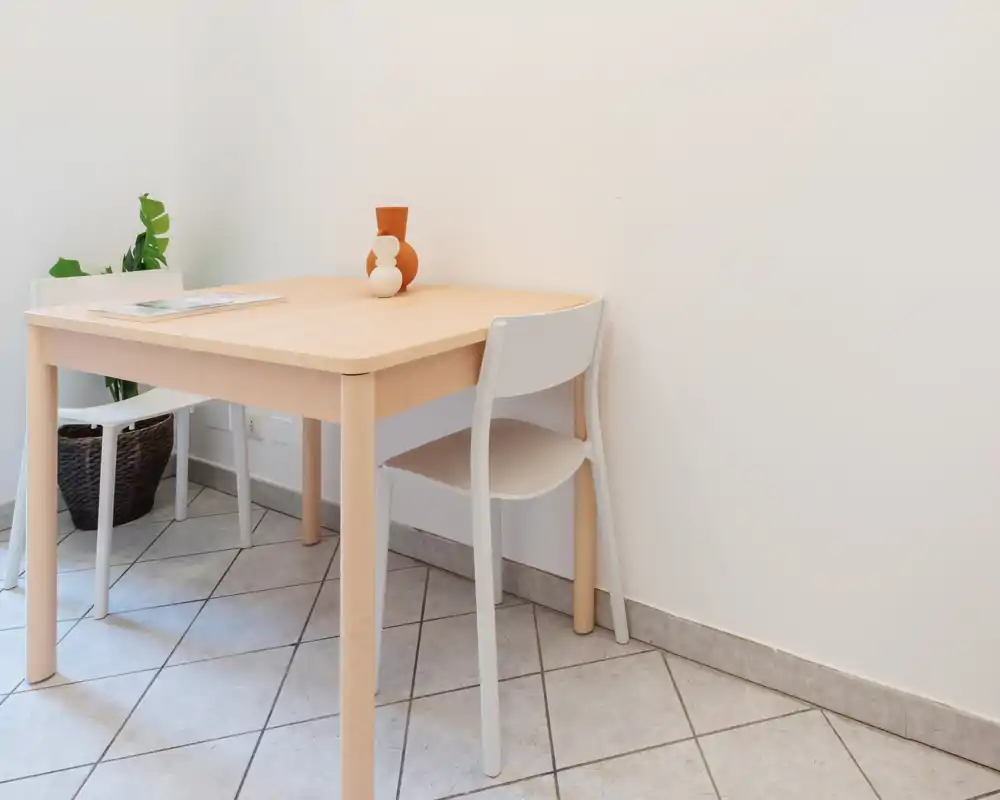 Angolo pranzo semplice e luminoso con tavolo in legno chiaro, due sedie bianche, un piccolo vaso decorativo e una pianta in vaso—ideale per pasti informali o un appartamento compatto.