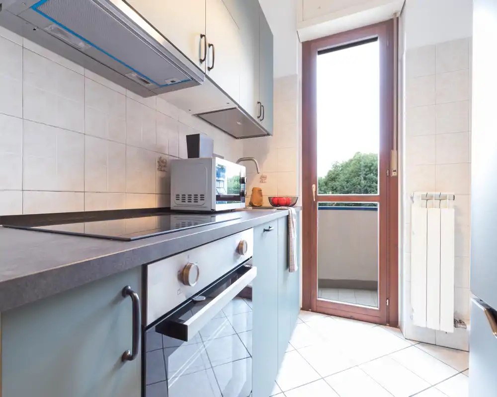 Cucina moderna ben illuminata con forno incorporato, piano a induzione e piccoli elettrodomestici sul piano; porta su un balcone che introduce molta luce naturale.