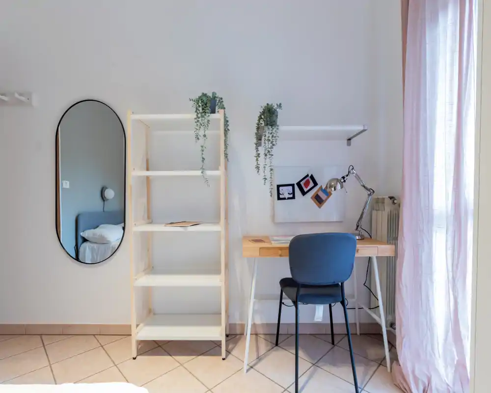 Coin chambre lumineux et minimaliste avec bureau, étagère et miroir reflétant une partie du lit. La lumière naturelle et les textiles doux apportent une ambiance calme pour travailler près du couchage.