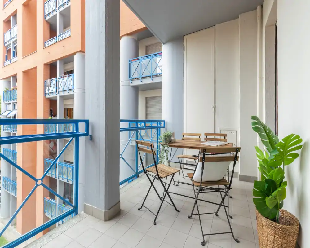 Balcon ensoleillé aménagé avec une petite table en bois et chaises pliantes, garde-corps bleu et plante en pot — idéal pour le café du matin ou un repas en plein air.