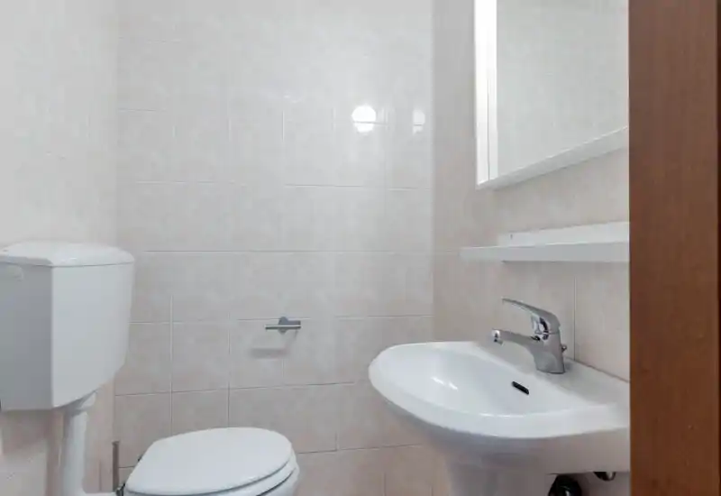 Compact, clean bathroom with a toilet, wall-mounted cistern and a pedestal sink beneath a mirror. Neutral tiled walls and simple chrome fixtures create a bright, functional space.