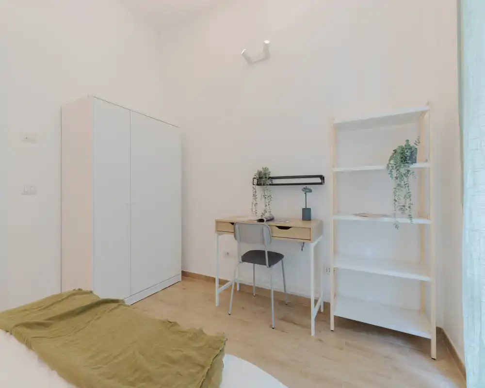 Camera luminosa e minimal con angolo del letto visibile, armadio bianco, una piccola scrivania in legno e scaffale con piante — ambiente pulito e arioso, ideale per una persona.