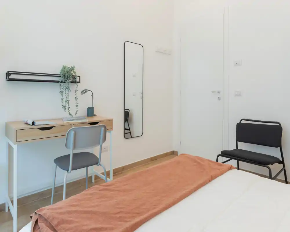 Camera da letto luminosa e minimal con letto ordinato, scrivania in legno e specchio a parete — ideale per un soggiorno tranquillo e adatto al lavoro.