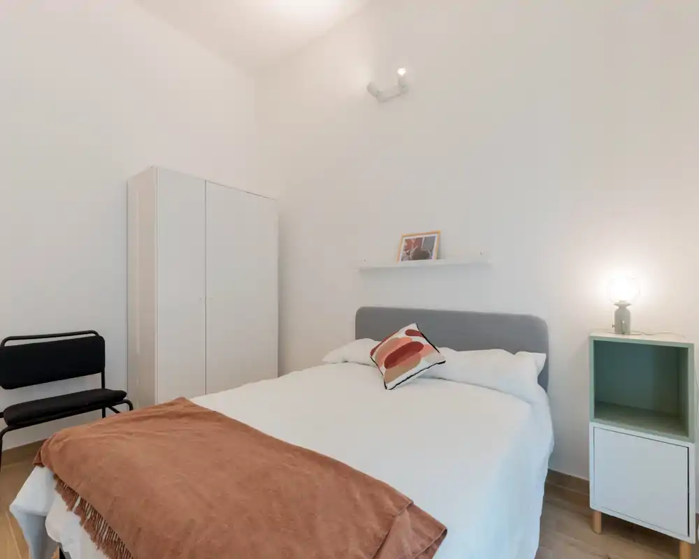 Camera da letto luminosa e minimal con letto matrimoniale ben rifatto, piccolo comodino con lampada, armadio e cuscino decorativo con plaid per un tocco accogliente.