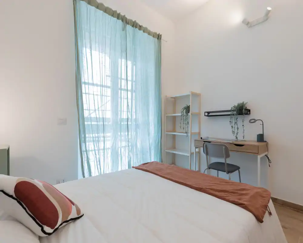 Camera da letto moderna e luminosa con letto ben rifatto, scrivania e scaffalatura vicino alla finestra; tende color turchese filtrano la luce naturale.