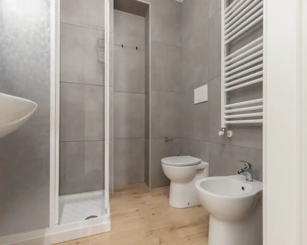 Bagno moderno e luminoso con cabina doccia, WC e bidet su pavimento effetto legno e grandi piastrelle grigie a parete.